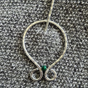 Peut inclure: Une épingle à châle argentée avec une texture martelée. L'épingle présente un design circulaire avec une perle verte et noire en bas et une épingle droite pour la fixer. L'épingle est présentée sur un fond texturé gris.