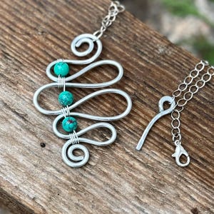 May include: A silver-toned necklace with a unique, flowing design. The pendant features a wavy pattern with three turquoise beads. The necklace has a delicate chain and a clasp for adjustable length. The jewelry is displayed on a wooden surface.