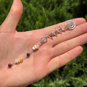 Op de afbeelding: Een zilverkleurig metalen haarspiraalaccessoire met een bungelende bedelketting. De ketting is versierd met kleine, onregelmatig gevormde kralen in tinten wit, crème, donkergrijs en een enkele ronde roodbruine kraal aan het uiteinde.