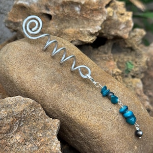 Op de afbeelding: Een zilverkleurig metalen haarspiraalaccessoire met een spiraalvormig ontwerp aan de bovenkant. Een kleine zilverkleurige karabijnsluiting is verbonden met een bungelende ketting van diverse onregelmatige, gepolijste blauwgroene stenen kralen en één grijze kraal.