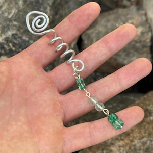 Op de afbeelding: Een haarspiraal van zilverkleurig metaal met een spiraalvormig bovendeel en een bungelende ketting. De ketting is versierd met twee doorschijnende groene kralen, een bleke matte kraal en een getextureerde groene kraal aan het uiteinde.