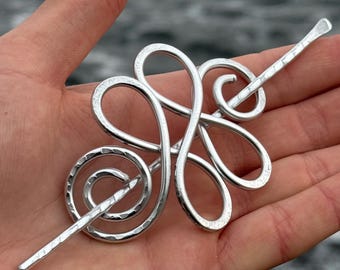 May include: A silver-colored hair pin with an intricate, abstract design. The pin features looping shapes and a spiral element, with a long, textured stick for insertion. The hair accessory is held in a hand.