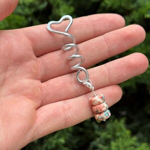 Op de afbeelding: Een zilverkleurige metalen haarspiraal met een hartvormige bovenkant en een bungelende bedel. De bedel bestaat uit drie kleine, ronde, veelkleurige kralen met een gemarmerd patroon in tinten rood, wit en blauwgroen.