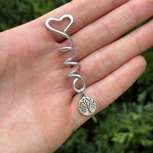 Op de afbeelding: Een zilverkleurige metalen haarspiraal met een hartvormige bovenkant en een kleine, ronde bungelende bedel met een gedetailleerd boomontwerp. De spiraal is gemaakt van een enkele draad die naar beneden krult om de ronde bedel vast te houden.
