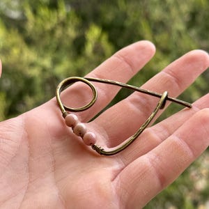 Op de afbeelding: Een handgemaakte haarspeld in bronskleur met een gebogen ontwerp. De speld heeft drie kleine, lichtroze kralen. De speld is ontworpen met een lusvormig uiteinde en een rechte speldsluiting. De achtergrond is een wazig groen.
