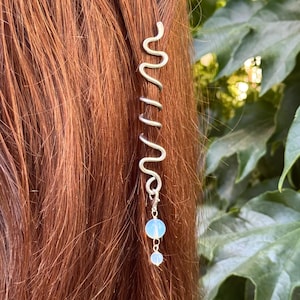 Op de afbeelding: Een zilverkleurige metalen haarspiraal met een golvend ontwerp dat om een haarlok heen draait. Het accessoire is afgewerkt met een klein zilveren kettinkje waaraan twee ronde, doorschijnende, lichtblauwe kralen hangen.