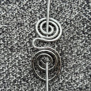 Puede incluir: Una horquilla plateada con un diseño en espiral. El pasador presenta dos espirales enrolladas conectadas por una línea curva, con un pasador recto que se extiende desde cada espiral. El fondo es una tela gris texturizada.