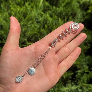 Op de afbeelding: Een zilverkleurig haarspiraalaccessoire met een gedraaide draadtop en een bungelende kettingbevestiging. De ketting bevat een kleine ronde bedel gegraveerd met de tekst "Beads of Life" en een gedetailleerde Hamsa-handbedel aan het uiteinde.