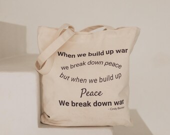 Może przedstawiać: Kremowa torba płócienna z dwoma paskami na ramię. Z przodu znajduje się czarny nadruk z tekstem: When we build up war we break down peace but when we build up Peace We break down war - Cindy Baxter.