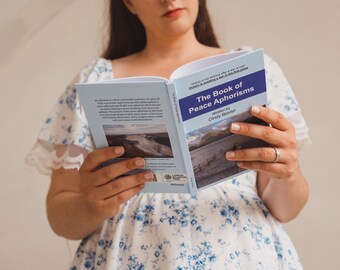 Może przedstawiać: Książka w miękkiej oprawie zatytułowana The Book of Peace Aphorisms autorstwa Cindy Baxter, z jasnoniebieską okładką, ciemnoniebieskim paskiem, białym tekstem i zdjęciem krajobrazu przedstawiającym puste pole. Książka jest otwarta, ukazując tył i przód.
