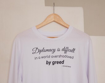 Może przedstawiać: Biała koszulka z długim rękawem wisząca na drewnianym wieszaku. Z przodu znajduje się czarny napis: Diplomacy is difficult in a world overshadowed by greed - Cindy Baxter. Koszulka ma okrągły dekolt i proste długie rękawy.