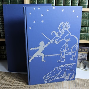 May include: A blue hardcover book featuring a silver foil illustration of a child dueling a pirate with a hook hand, with a crocodile below and stars above. A second plain blue book stands behind it, both with a smooth, matte-textured finish.