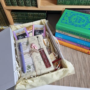 May include: A gift box containing a wrapped book, a purple pen, a dark red metal bookmark, Twinings Earl Grey tea bags, and a small biscuit. Beside the box is a stack of five colourful hardcover books titled The Tenant of Wildfell Hall, The Professor, Villette, Agnes Grey, and Jane Eyre.