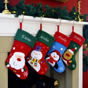 May include: Four Christmas stockings hang from a mantelpiece. Each stocking has a different design: Santa Claus, a snowman, a penguin, and a reindeer. Names are embroidered on each stocking, with a red loop for hanging.