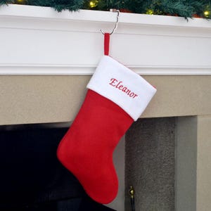 May include: A festive red Christmas stocking with a white cuff, embroidered with the name "Eleanor" in red. It hangs from a silver hook on a white mantelpiece, with decorative greenery and lights above.