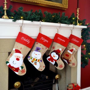 May include: Four festive Christmas stockings suspended from a mantelpiece. Each stocking has a red cuff with a name embroidered in white: Daddy, Jürgen, Alexandra, and Oliver. The stockings are decorated with festive characters.