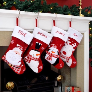 May include: Four red Christmas stockings with white cuffs hang above a fireplace. Each stocking features a festive character: Santa, a snowman, a penguin, and a reindeer. Names are embroidered on the cuffs.