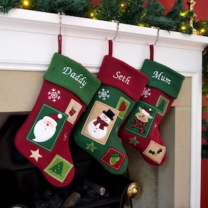 May include: Three Christmas stockings hang from a mantelpiece. Each stocking is red and green with embroidered details and the names "Daddy", "Seth", and "Mum". The stockings feature images of Santa, a snowman, and a reindeer.