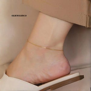 Op de afbeelding: Een delicate gouden enkelband wordt om een enkel gedragen. De enkelband is een dunne, eenvoudige ketting. De voet rust op een witte schoen met een bruine zool. De tekst "OZJEWELRYCO" is zichtbaar.