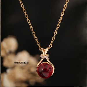 May include: A gold-colored necklace with a pendant featuring a round, deep red gemstone. The pendant is held in a gold setting with a small, sparkling accent. The chain is delicate and the background is dark.