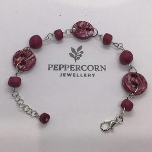 May include: A burgundy and white marbled button bracelet with silver chain links and a lobster clasp. The bracelet features round and oval beads. The text "PEPPERCORN JEWELLERY" is printed on the background.