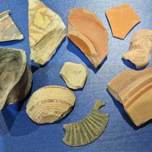 Puede incluir: Una colección de fragmentos de cerámica antigua de diversas formas y tamaños, con colores descoloridos como beige, naranja, gris y verde. Algunas piezas muestran restos de diseños y patrones pintados, lo que sugiere su importancia histórica.