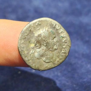 Könnte beinhalten: Eine verwitterte, runde Münze mit dem Profil eines Kopfes. Die Münze hat ein angelaufenes, metallisches Aussehen mit sichtbaren Inschriften und Details. Die Münze ist wahrscheinlich ein antikes oder historisches Artefakt.