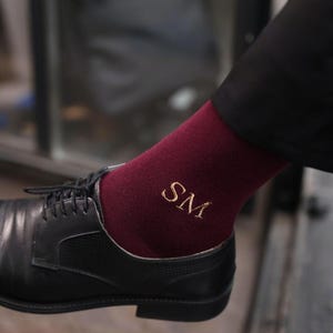 Puede incluir: Calcetines burdeos con las iniciales doradas "SM" bordadas, visibles sobre un zapato de vestir de cuero negro. Los calcetines son de color liso y las iniciales están en una fuente sencilla. El zapato tiene un diseño clásico con cordones.