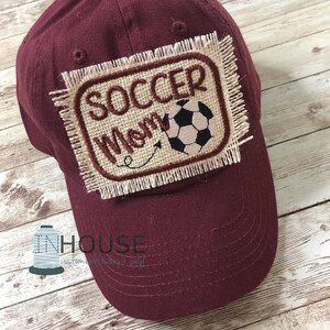 May include: Burgundy baseball cap with a beige patch that says "Soccer Mom" with a soccer ball graphic.