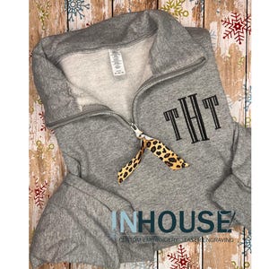 May include: A gray sweatshirt with a zipper and a black embroidered monogram "THT". A leopard print ribbon is tied around the zipper pull. The sweatshirt is on a wooden background.