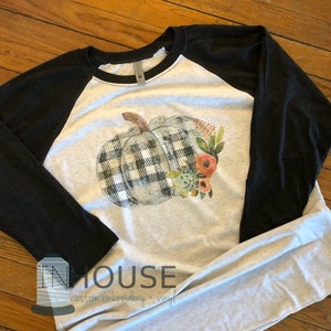May include: A white and black checkered pumpkin with floral accents on a gray and black baseball tee shirt. The shirt has the text "IN HOUSE custom embroidery + vinyl" on the bottom left corner.