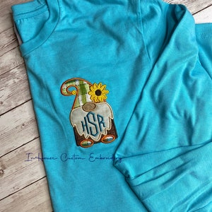 May include: A turquoise long-sleeved t-shirt with a gnome embroidered on the front. The gnome is wearing a plaid hat with a sunflower on top and has a monogram "HSR" on its chest.