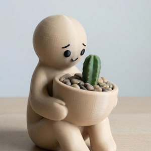 May include: A beige figurine of a person holding a small, round pot. The pot contains a green cactus and small pebbles. The figurine has a simple face with black eyes and a smile. The overall design is minimalist and modern.