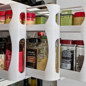 May include: White, pull-out spice rack with multiple shelves. The shelves hold various spice jars with red, black, and green lids. Some jars have labels like "Ground Ginger" and "Celery".