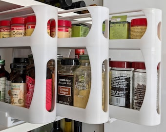 May include: White, pull-out spice rack with multiple shelves. The shelves hold various spice jars with red, black, and green lids. Some jars have labels like "Ground Ginger" and "Celery".