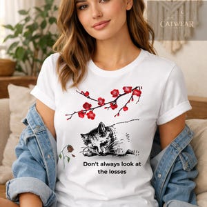 Può includere: T-shirt bianca con un'illustrazione in bianco e nero di un gatto che dorme e un ramo floreale rosso. Il testo "Don't always look at the losses" è stampato sotto.