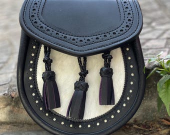 May include: A black leather sporran with a white fur front and three black tassels with purple detailing. The sporran features a decorative perforated flap and a circular design with white stitching.