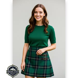 May include: A woman wearing a green short-sleeved top and a green plaid skirt with black and white accents. The skirt has two leather buckle closures. The Highland Legacy logo is in the bottom left corner.