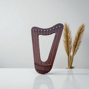 Pode incluir: Uma pequena harpa de madeira marrom escuro com pinos de afinação e cordas prateadas. A harpa está sobre um fundo branco com grama ornamental seca. O instrumento tem um topo curvo e uma base arredondada.