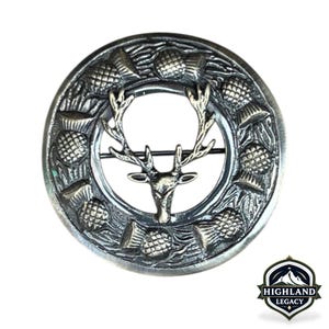 May include: A silver-toned brooch with a circular design. The brooch features a stag's head in the center, surrounded by a ring of thistles. The brooch has a pin on the back for secure fastening. The Highland Legacy logo is in the bottom right corner.