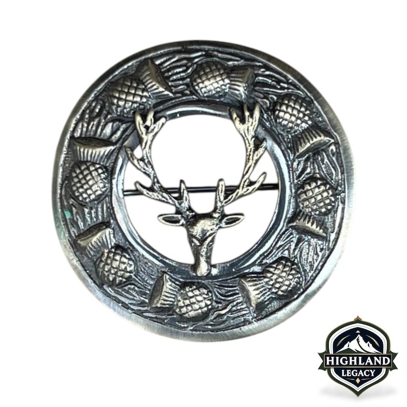 May include: A silver-toned brooch with a circular design. The brooch features a stag's head in the center, surrounded by a ring of thistles. The brooch has a pin on the back for secure fastening. The Highland Legacy logo is in the bottom right corner.