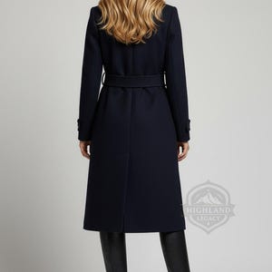 May include: A navy blue coat with a belt, long sleeves, and a classic design. The coat is knee-length and worn with black boots. The Highland Legacy logo is visible on the lower right.