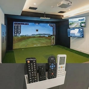 May include: A golf simulator room featuring a large screen showing a golf course. A white holder contains several remotes. Additional screens display course information. The room has green turf flooring and a projector mounted on the ceiling.