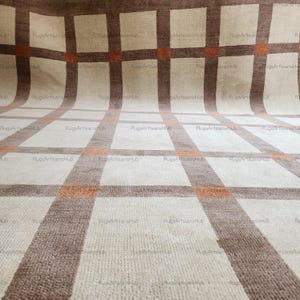 Può includere: Un tappeto a quadri beige e marrone con una superficie strutturata. Il tappeto presenta un motivo a griglia con linee marroni e arancioni alternate. Il tappeto è realizzato in un materiale morbido ed è adatto per un soggiorno o una camera da letto.