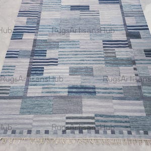 Puede incluir: Una alfombra rectangular con un patrón geométrico en tonos azules, grises y blancos. El diseño presenta un mosaico de rectángulos y rayas. La alfombra tiene un borde con flecos y es adecuada para la decoración del hogar.