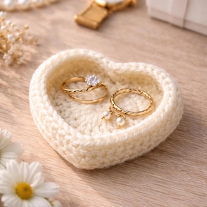 May include: A heart-shaped, cream-colored, crocheted ring dish holding two gold rings. One ring has a large, clear stone, and the other has two small pearl charms. The dish sits on a light-colored wooden surface, with flowers and a watch in the background.