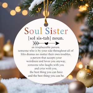 Puede incluir: Adorno navideño redondo y blanco con las palabras "Soul Sister" en texto colorido. El adorno incluye una definición de "Soul Sister" y cuelga de un hilo dorado, con decoraciones navideñas en el fondo.