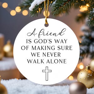 May include: A white, round Christmas ornament with the text "A friend is God's way of making sure we never walk alone" and a cross. The ornament hangs from a gold string, with a blurred background of Christmas decorations.