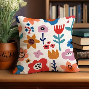 May include: Square throw pillow with a white background and a colorful floral pattern. The flowers are in shades of orange, red, blue, pink, and yellow. The pillow is likely made of a soft fabric and is designed for decorative use.