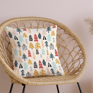 Peut inclure: Un coussin carré avec un motif répété d'arbres de Noël stylisés en rouge, noir, or et bleu sur fond crème. Le coussin est posé sur une chaise en osier.